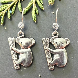 May include: Silver-toned koala bear earrings. Each earring features a koala clinging to a tree branch, with a sparkling clear gem at the top. The earrings are set against a dark gray background with green foliage.