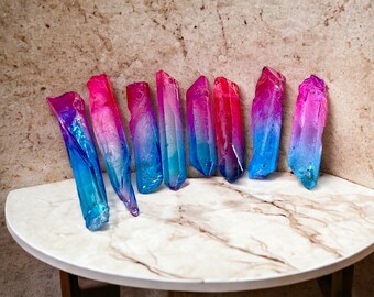 Könnte beinhalten: Eine Sammlung von acht schillernden Kristallspitzen mit einem Farbverlauf von Pink zu Blau. Die Spitzen sind in einem Halbkreis auf einer weißen Marmorfläche angeordnet.