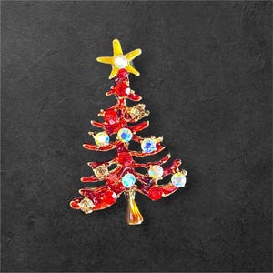 May include: A Christmas tree brooch with a yellow star on top. The red tree is adorned with colorful, iridescent jewels. The brooch is set against a dark background.