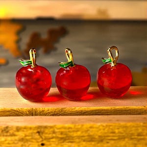 May include: Three small, red glass apple ornaments with gold stems and green leaves. The apples are displayed on a wooden surface, with a blurred background. The ornaments have a shiny, transparent appearance.