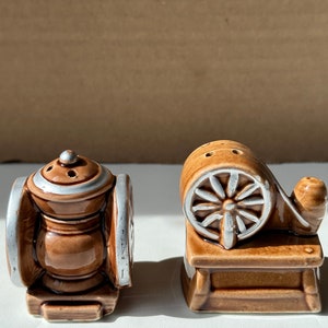 May include: A set of two brown ceramic salt and pepper shakers. The shakers are shaped like old-fashioned machinery. One shaker has a wheel and the other has a lantern shape.