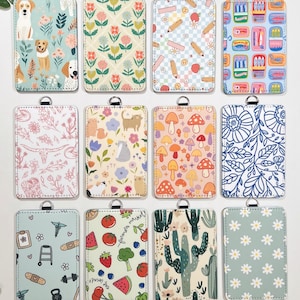 May include: A collection of rectangular ID badge holders with various patterns. Designs include dogs, flowers, pencils, food, cats, mushrooms, and cacti. Each holder has a silver-colored ring at the top for attaching a lanyard.