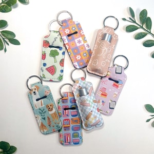 May include: A collection of seven keychains, each featuring a different patterned design. The patterns include floral, dog, book, and fruit motifs. Each keychain has a silver ring and a holder for a lip balm tube.