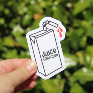 May include: A white sticker featuring a juice box design with a straw and red drips. The text "Juice Collector" is printed on the front of the juice box. The sticker is held in a hand against a blurred green background.