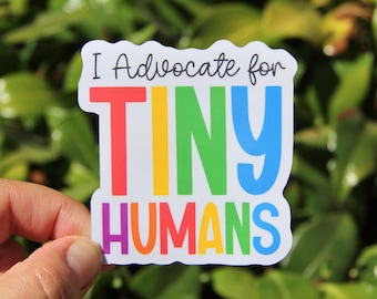 May include: A white sticker with the text "I Advocate for TINY HUMANS" in a colorful rainbow design. The sticker is held by a hand against a backdrop of green foliage.