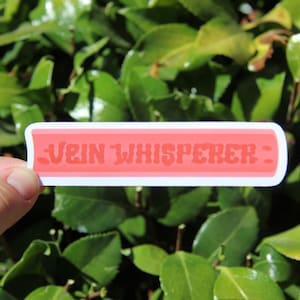 May include: A rectangular sticker with the text "VEIN WHISPERER" in a coral-colored, stylized font. The text is set against a coral background with a white border. The sticker is held against a backdrop of green leaves.