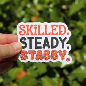 May include: A white sticker with the words "SKILLED. STEADY. STABBY." in a bold, retro font. The word "SKILLED" is in red, "STEADY" is in black, and "STABBY" is in orange. The sticker is held against a blurred green background.