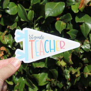 May include: A white pennant-shaped sticker with the words "1st grade TEACHER" in pastel colors. The sticker has light blue accents and is held against a backdrop of green leaves.