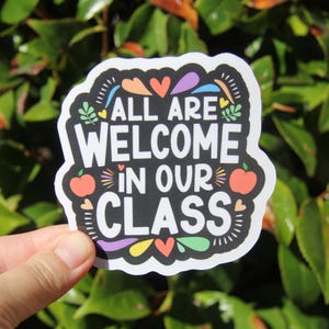 May include: A sticker with the text "ALL ARE WELCOME IN OUR CLASS" in white letters on a black background. The sticker has colorful accents, including hearts, apples, and leaves. The sticker is held in front of a green leafy background.