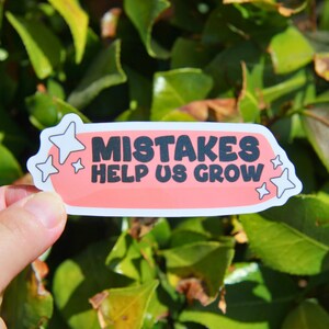 May include: A sticker with the text "MISTAKES HELP US GROW" in black letters on a pink rectangular background. The sticker has a white border and star-shaped accents. The sticker is held in front of a background of green leaves.