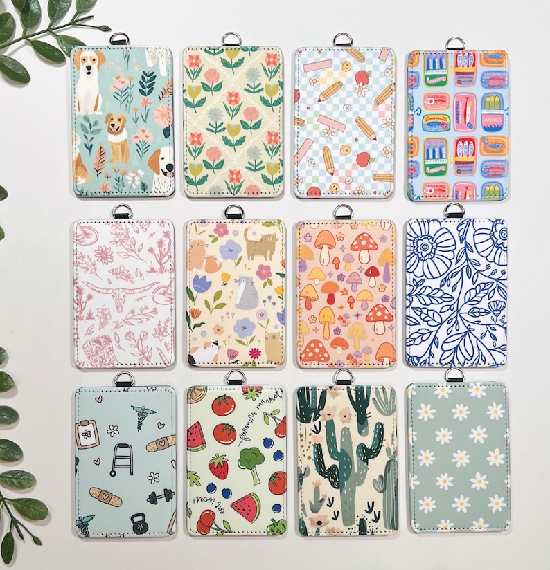 May include: A collection of rectangular ID badge holders with various patterns. Designs include dogs, flowers, pencils, food, cats, mushrooms, and cacti. Each holder has a silver-colored ring at the top for attaching a lanyard.