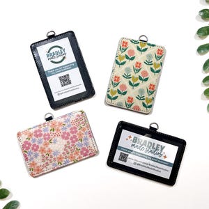 Op de afbeelding: Vier ID-badgehouders met verschillende ontwerpen. Twee zwarte houders met doorzichtige vensters tonen tekst en een QR-code. De andere twee hebben bloempatronen in roze, geel en blauw. Elk heeft een zilveren ring.