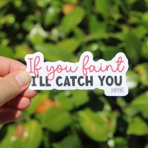 May include: A white sticker with the text "If you faint I'll catch you" in a mix of red and black fonts. The word "maybe" is in a smaller font at the bottom. The sticker is held in a hand against a blurred green background.