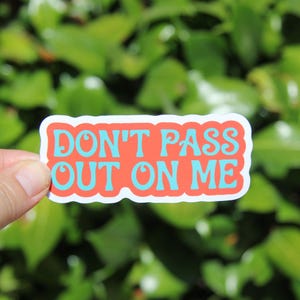 May include: A rectangular sticker with the text "DON'T PASS OUT ON ME" in turquoise letters on a red background, outlined in white. The sticker is held against a blurred green leafy background.