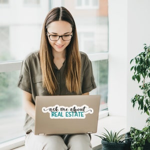 Puede incluir: Una mujer con gafas se sienta frente a una ventana, usando una computadora port&aacute;til. La computadora port&aacute;til tiene una pegatina que dice "ask me about REAL ESTATE".