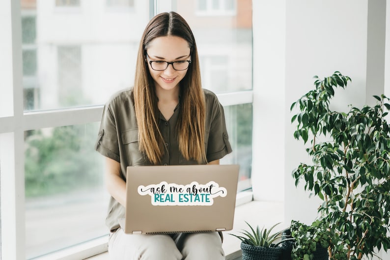 Puede incluir: Una mujer con gafas se sienta frente a una ventana, usando una computadora port&aacute;til. La computadora port&aacute;til tiene una pegatina que dice "ask me about REAL ESTATE".