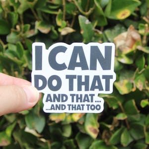 May include: A white sticker with the text "I CAN DO THAT" in a bold, dark gray font. Below, the text continues with "AND THAT... ...AND THAT TOO" in a smaller font. The sticker is held against a backdrop of green foliage.