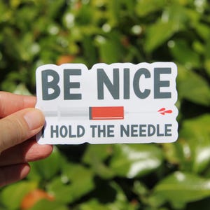 May include: A white sticker with the text "BE NICE" in gray block letters above "HOLD THE NEEDLE" in white letters. A syringe graphic is in the center with a red section and a red droplet design.