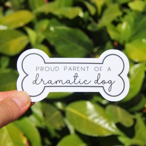May include: A white sticker in the shape of a dog bone with the text "PROUD PARENT OF A dramatic dog" in black script. The sticker is held against a background of green leaves.