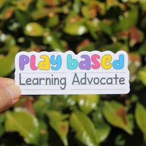 May include: A white sticker with the words "Play Based Learning Advocate" in colorful bubble letters and a handwritten font. The sticker is held against a backdrop of green foliage.