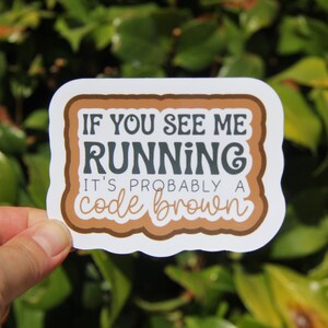May include: A white sticker with a brown border and the text "IF YOU SEE ME RUNNING IT'S PROBABLY A code brown" in brown and gray lettering. The sticker is held in front of a green leafy background.