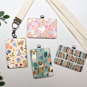Op de afbeelding: Vier rechthoekige ID-badgehouders met verschillende ontwerpen. E&eacute;n heeft een bloemenpatroon, een andere heeft katten, een derde heeft honden en de laatste heeft een boekenplankenpatroon. Ze zijn bevestigd aan een cr&egrave;mekleurig koord.