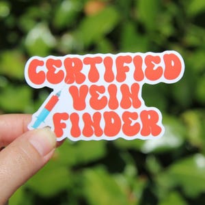 May include: A white sticker with the words "CERTIFIED VEIN FINDER" in orange bubble letters. A small syringe with a red and blue design is on the left side of the sticker. The sticker is held in front of a green background.