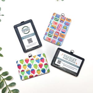 Op de afbeelding: Vier ID-badgehouders met verschillende ontwerpen. Twee zijn zwart met een doorzichtig venster en de tekst "BRADLEY made studios". De andere twee hebben kleurrijke patronen: aardbeien en een sardine-ontwerp. Elk heeft een zilveren ring.