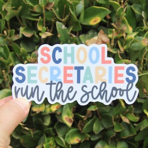 May include: A colorful sticker with the text "SCHOOL SECRETARIES run the school." The text is in various colors, including blue, orange, and green, against a white background. The sticker is held in front of a green leafy background.