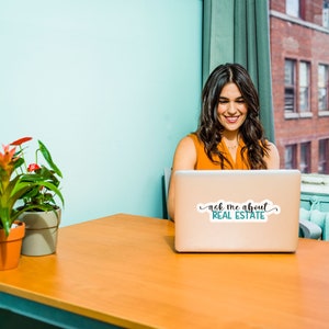 Puede incluir: Una mujer se sienta en un escritorio frente a un ordenador port&aacute;til. El port&aacute;til tiene una pegatina que dice "ask me about real estate". Hay dos plantas en macetas en el escritorio.