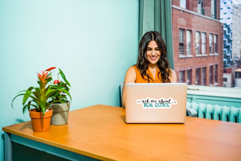 Puede incluir: Una mujer se sienta en un escritorio frente a un ordenador port&aacute;til. El port&aacute;til tiene una pegatina que dice "ask me about real estate". Hay dos plantas en macetas en el escritorio.