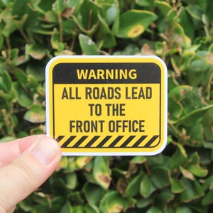 May include: A yellow and black sticker with the text "WARNING ALL ROADS LEAD TO THE FRONT OFFICE." The sticker has a black border and a diagonal striped design at the bottom. The sticker is held in front of a green leafy background.