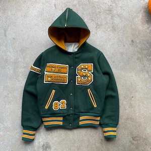 May include: A dark green varsity jacket with a hooded collar and gold detailing. The jacket displays patches with the text "WESCO CHAMPS 80" and "NW DISTRICT CHAMPS 80". The number "82" is also visible. The jacket has a button-up front and striped cuffs.