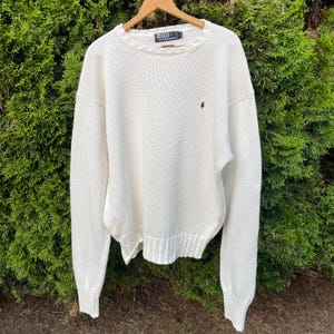 May include: A cream-colored long-sleeve knit sweater with a crew neckline and ribbed cuffs and hem. The garment features a small dark embroidered logo on the left chest and a Polo by Ralph Lauren label at the inner neck collar area.