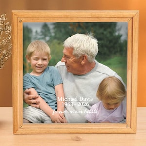 May include: A square wooden picture frame containing a photograph of an older adult and two children. The text Michael Davis 1963-2026 forever in our hearts is printed in white on the image. The frame has a light brown finish and a simple border.