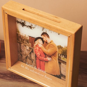 May include: A wooden shadow box with a slot on top, displaying a photograph of a couple. The photo is framed with the text "Lori & Sean let the adventure begin." A small, orange candle sits to the left.