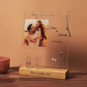May include: A clear acrylic jewelry organizer with a castle-shaped design. It features a photo frame, a necklace holder, a ring holder, and a section with small holes for earrings. The base is made of wood and has the text "Your Custom Text" engraved on it. The photo frame shows a couple smiling at each other. The text "James Amanda 01.20.2024" is engraved on the acrylic.
