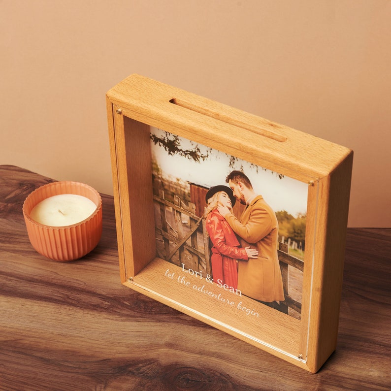 May include: A wooden shadow box with a slot on top, displaying a photo of a couple. The photo box has the text "Lori & Sean let the adventure begin". A small, peach-colored candle sits to the left.