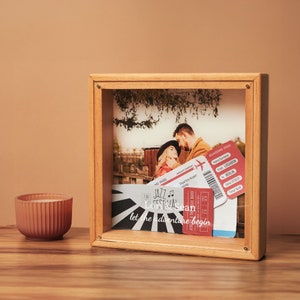 May include: A wooden shadow box frame displaying a photograph of a couple, along with travel tickets. The tickets are red and white, with text that reads "Lissabon to Berlin". The frame also includes the text "let the adventure begin".