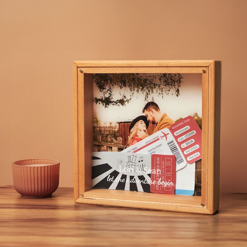 May include: A wooden shadow box frame displaying a photograph of a couple, along with travel tickets. The tickets are red and white, with text that reads "Lissabon to Berlin". The frame also includes the text "let the adventure begin".