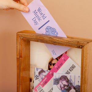 May include: A wooden shadow box with a collection of mementos. A hand inserts a light purple card with "World Music Day" printed on it. Other items include a pink ticket and a photograph.