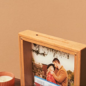 May include: A light-colored wooden shadow box with a slot on top, displaying a photograph of a couple. The photo box also contains a boarding pass. The box is made of light-colored wood and has a simple, clean design.