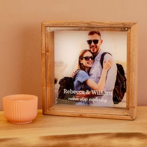 May include: A wooden shadow box frame with a photograph of a couple, with the text "Rebecca & William never stop exploring." A peach-colored, ribbed candle holder sits to the left of the frame.