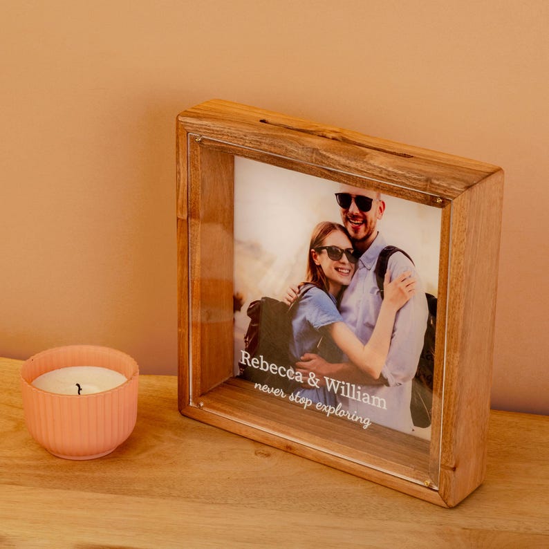 May include: A wooden photo frame with a clear front displays a couple's photo. The frame has the text "Rebecca & William never stop exploring." A small, pink candle sits to the left of the frame.
