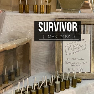 May include: A display of handmade candles in brass shell casings. The candles are arranged on a white tablecloth, with the word "SURVIVOR" and "MAN-DLES" in the image. A sign indicates the candles are made with beeswax.