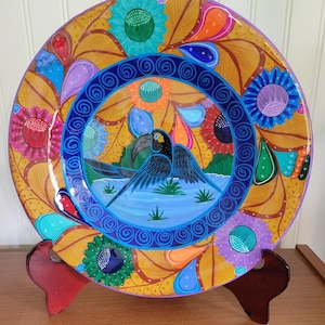 May include: A colorful ceramic plate with a blue and black parrot design. The plate has a yellow background with blue, green, purple, and orange accents. The parrot is perched on a branch with a blue and green background.