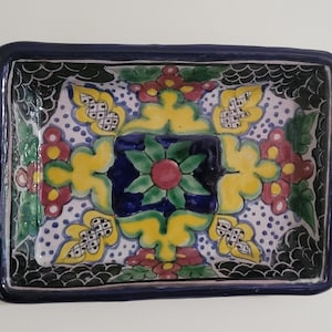 May include: Rectangular ceramic dish with a dark blue border. The interior features a central floral design in blue, green, yellow, and red. The background is white with small blue dots. The dish is likely handmade.
