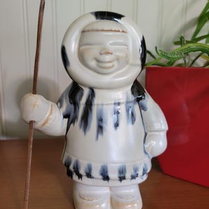May include: A ceramic figurine of a person wearing a hooded parka and holding a stick. The figurine is painted in white, black, and blue.