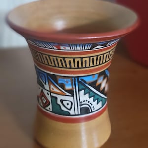 May include: A small, brown ceramic vase with a geometric pattern in shades of blue, green, red, and black. The vase has a wide mouth and a narrow base.