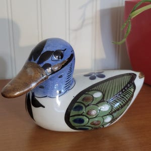 May include: A ceramic duck figurine with a blue head, brown beak, and white body. The duck has black and green wing details and blue floral accents. The figurine is sitting on a wooden surface.